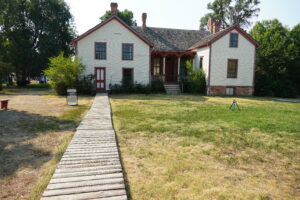 A wooden pathway, flanked by grass, leads to a white house with red trim.