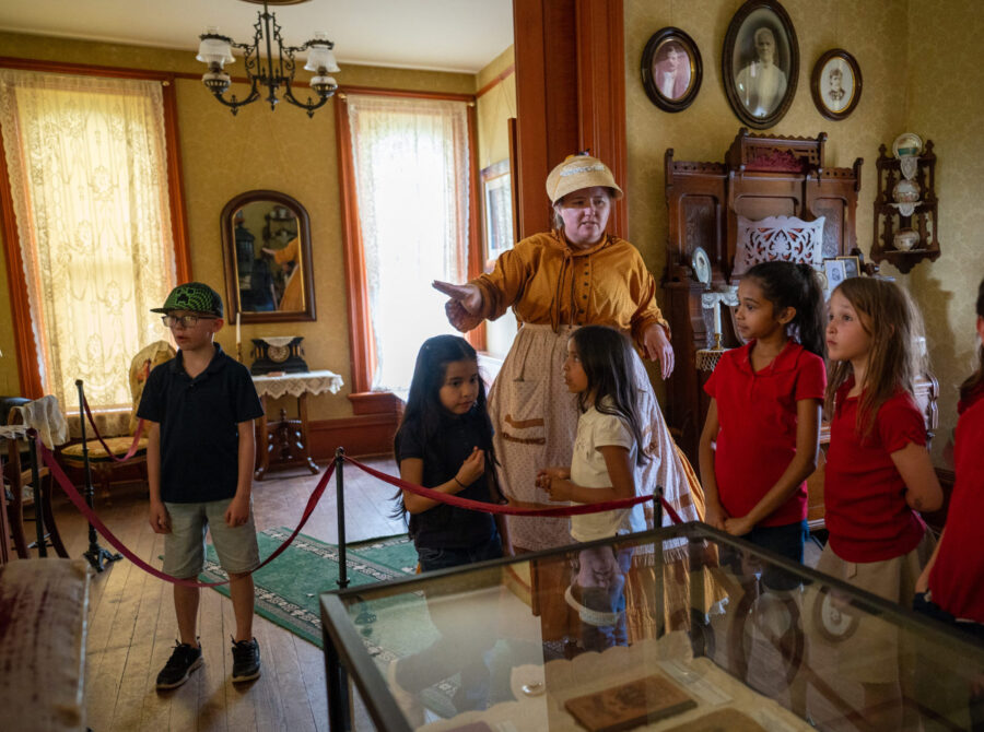A woman in historical costume guides children in a period room.