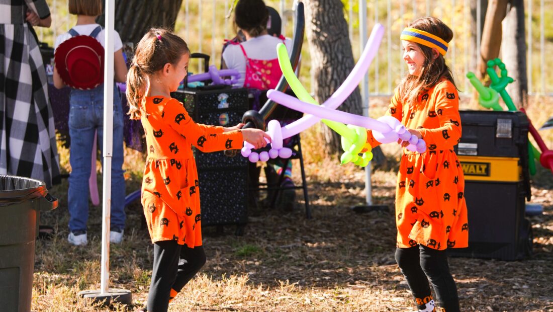 Two girls in matching orange dresses play with balloon swords