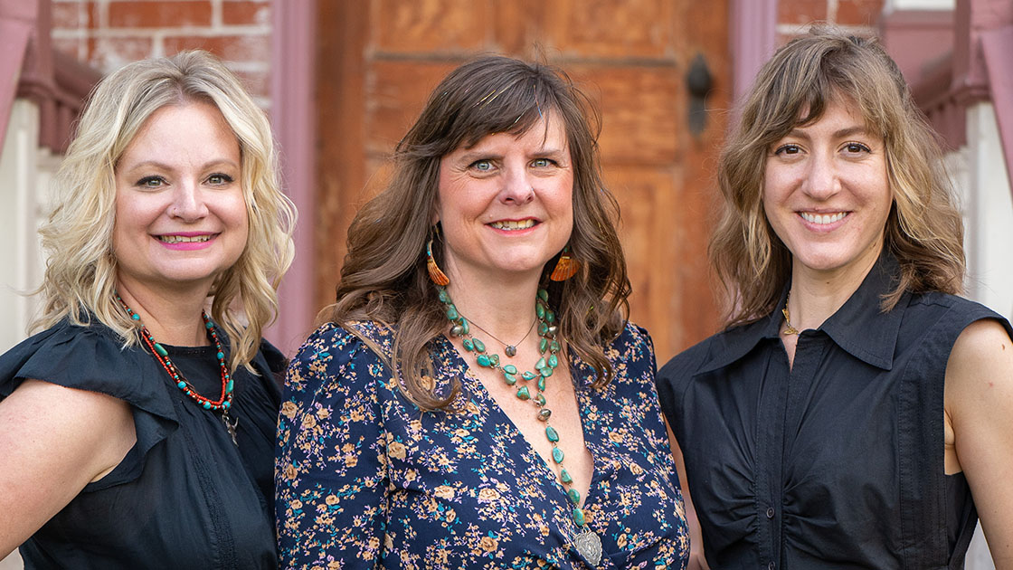 Three women smiling, standing in front of a door.