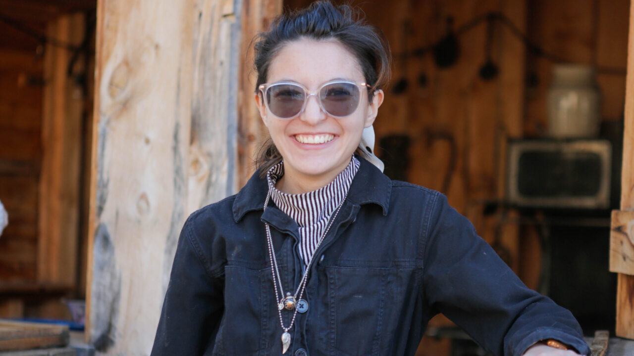 Person smiling outdoors, wearing sunglasses and a black jacket