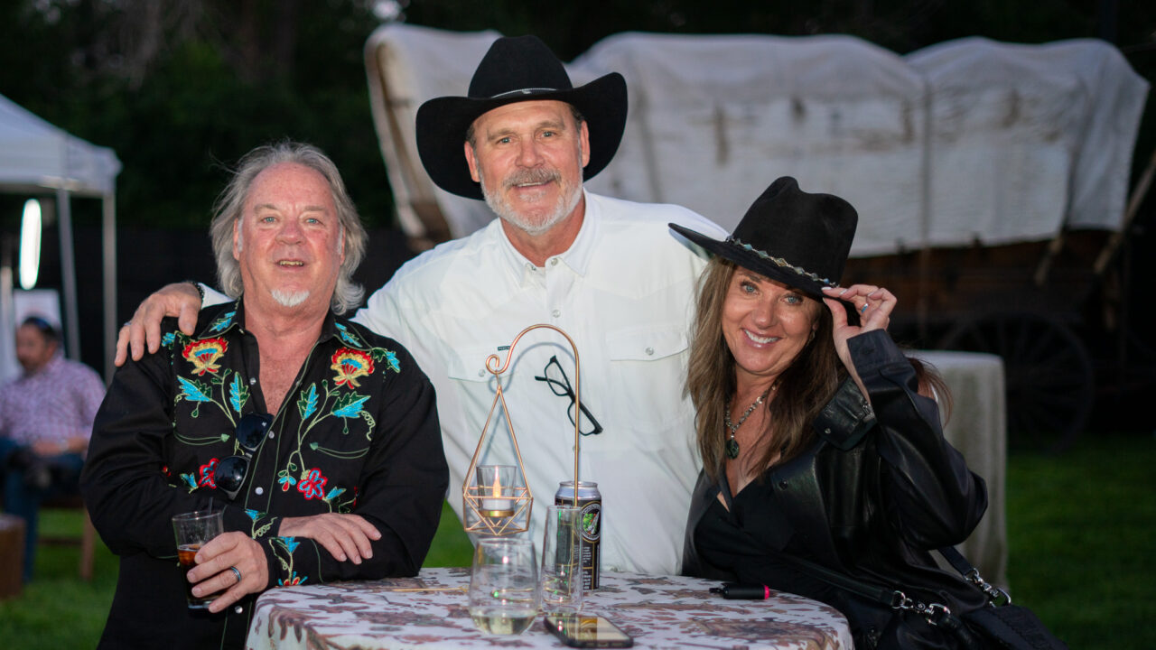 Three people wearing cowboy hats smiling at an outdoor event, with a table and covered wagon in the background