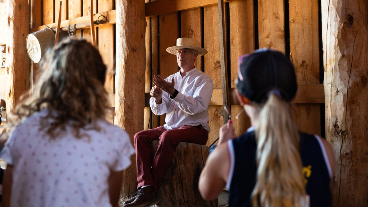 Man in straw hat explaining something to children inside a rustic wooden barn
