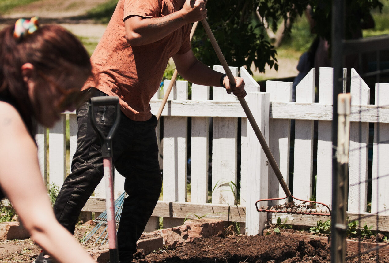 Man raking soil near white fence, woman digging with shovel