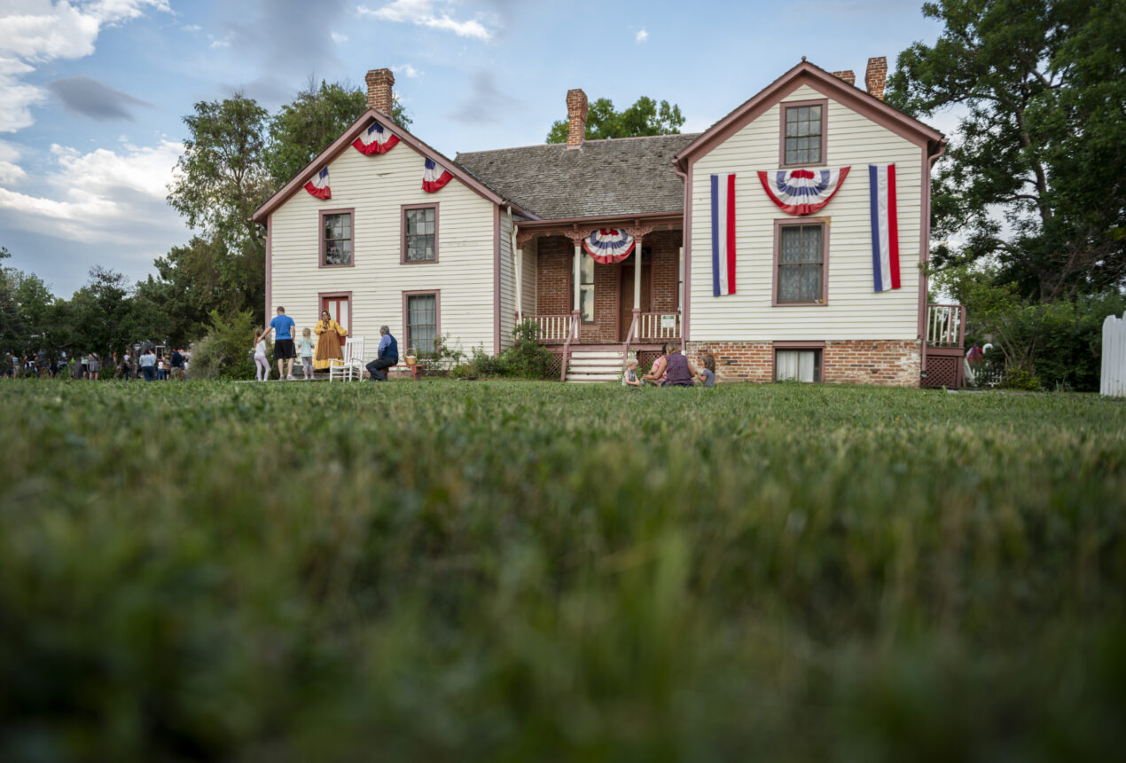 Historic house with red, white, and blue bunting; people gathered on porch and lawn