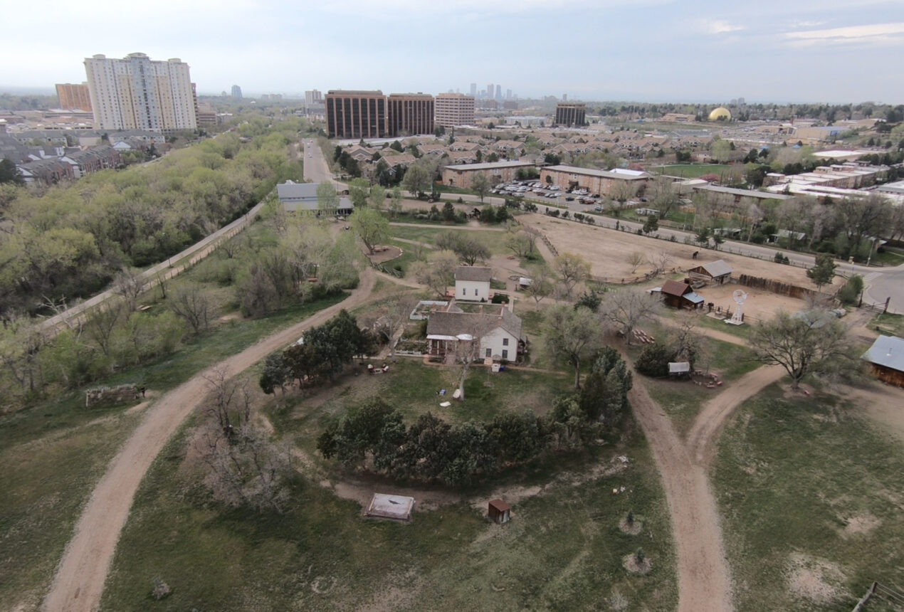 Aerial view of a rural area with houses, trees, and dirt paths, set against a backdrop of urban buildings in the distance