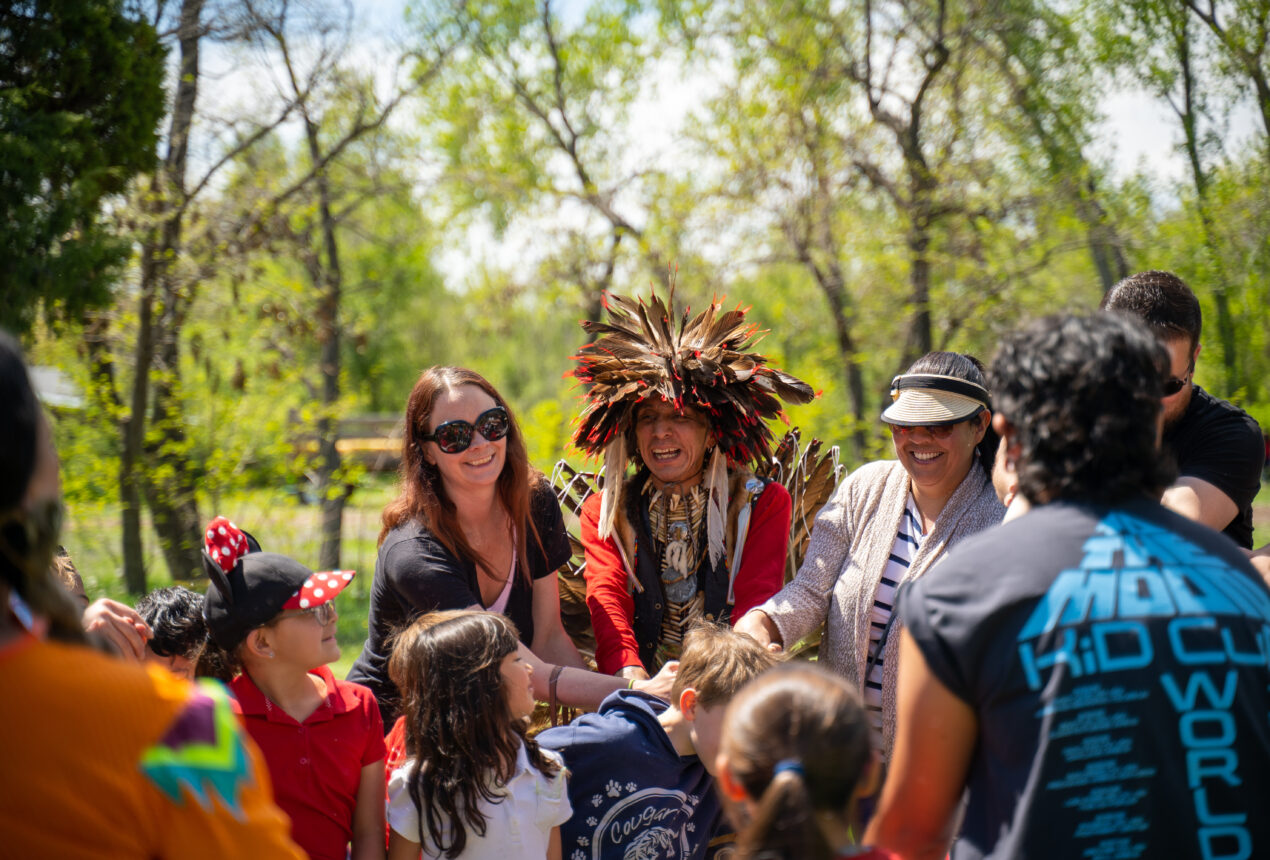 A group at Four Mile Park encircles a person in a feathered headdress, smiling and engaging together in an outdoor setting