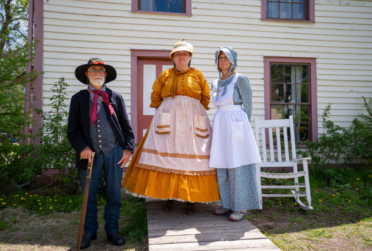 Three people wearing historical costumes in front of a white wooden house