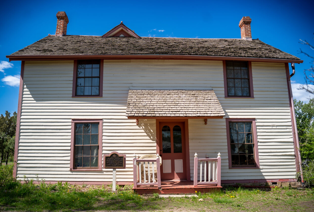 Historic two-story house with wooden siding, red trim, and a plaque near the entrance in a park setting