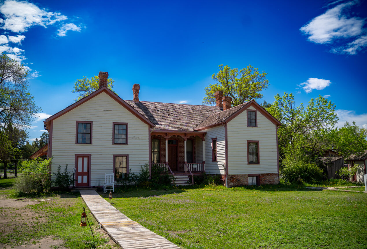 Historic white house with red trim, brick chimney, wooden walkway, surrounded by green lawn and trees under a blue sky