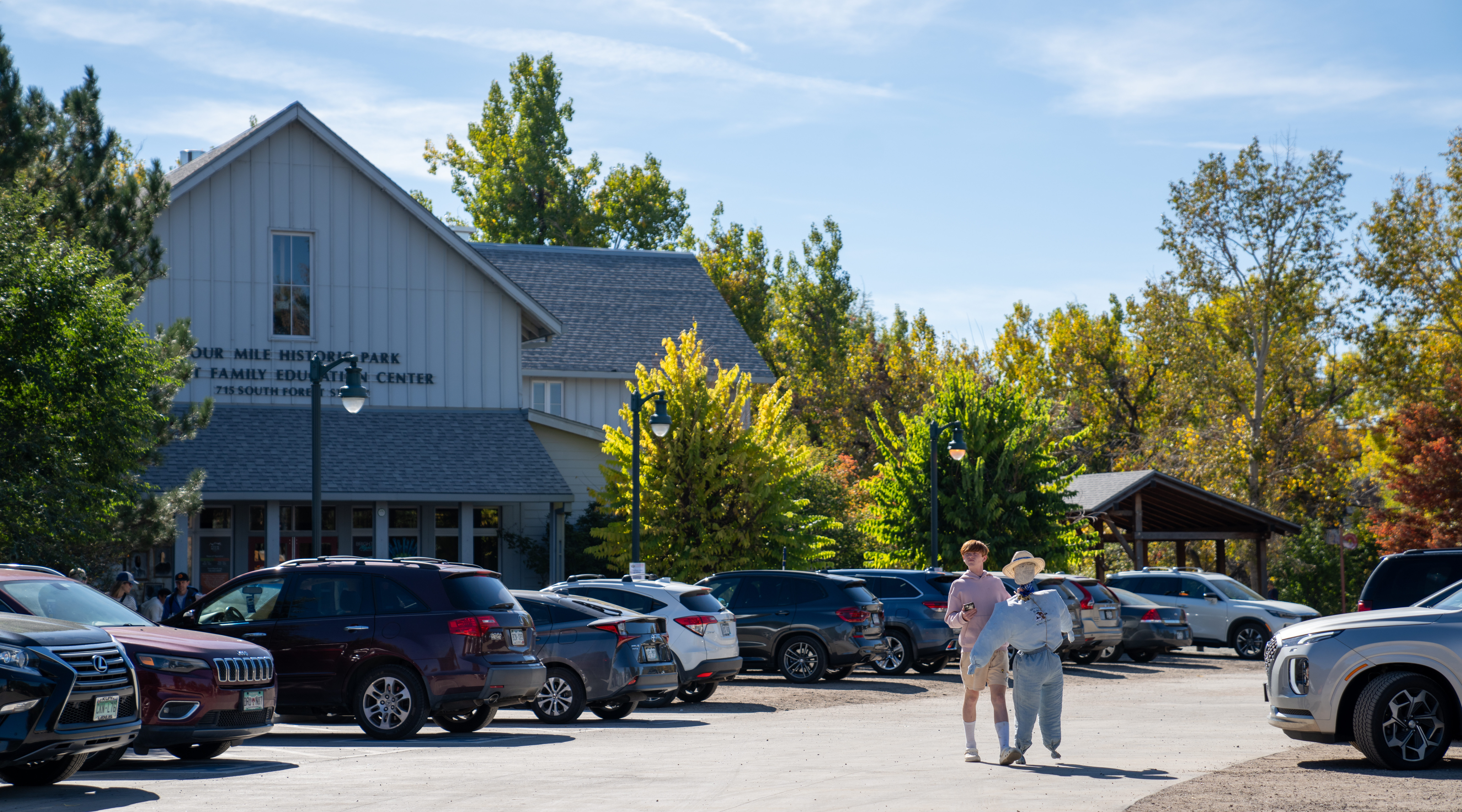 Person walking through a parking lot, cars parked under blue sky, outside Four Mile Historic Park Building