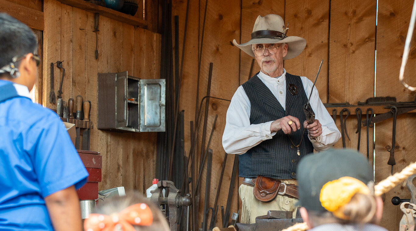 Older man in hat demonstrating a tool to an audience in a workshop
