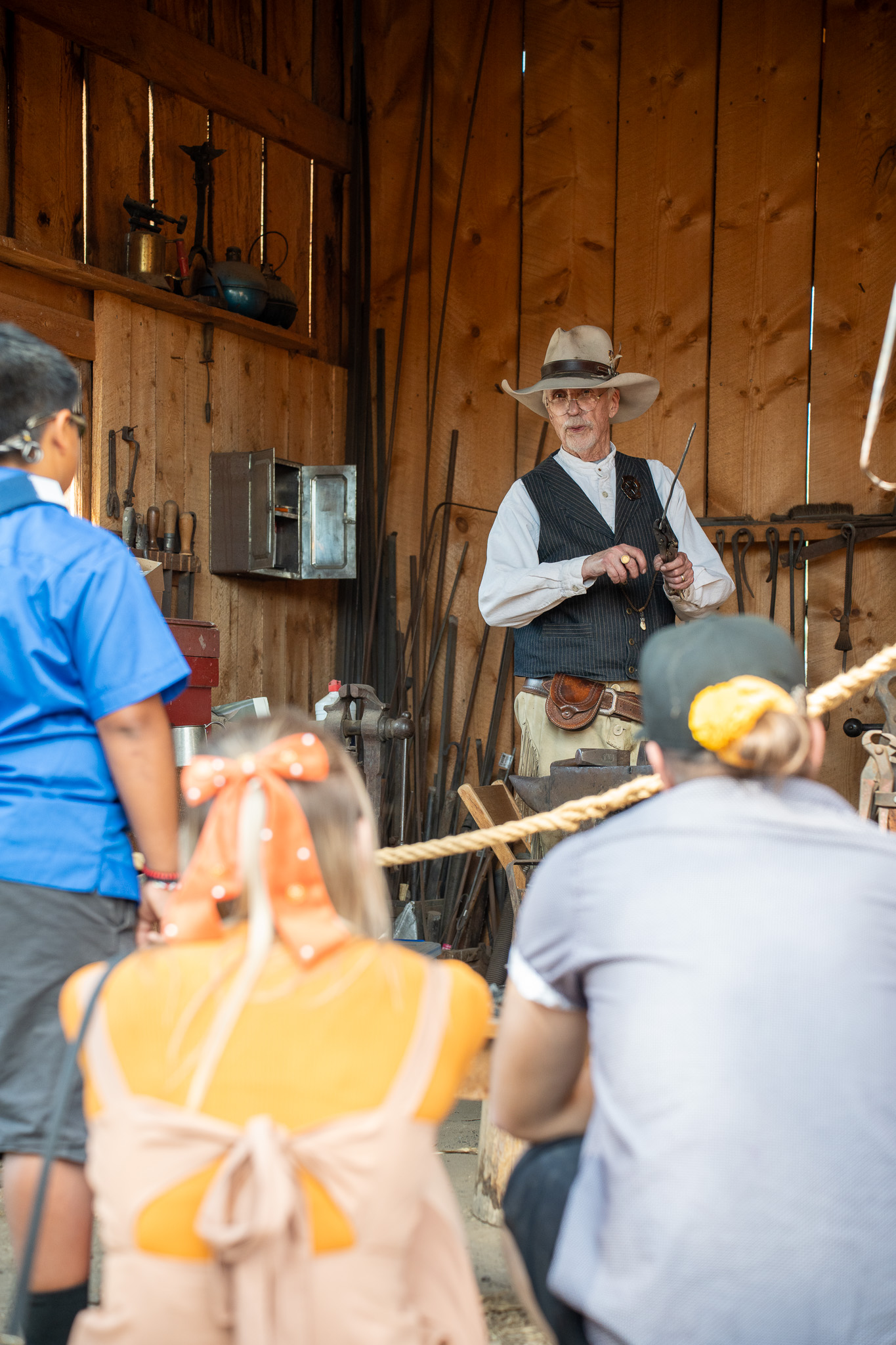 Person in hat demonstrating blacksmithing to attentive audience inside wooden workshop