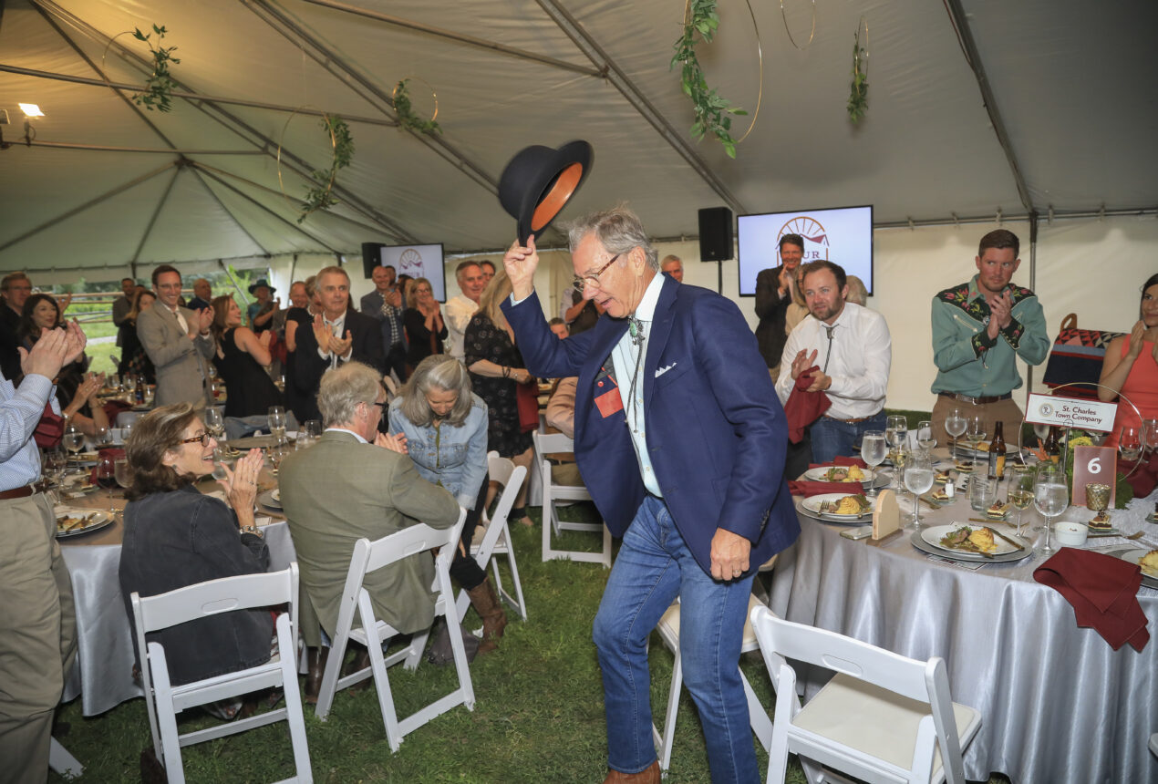 Man in blue jacket tips hat amid applause under a tent