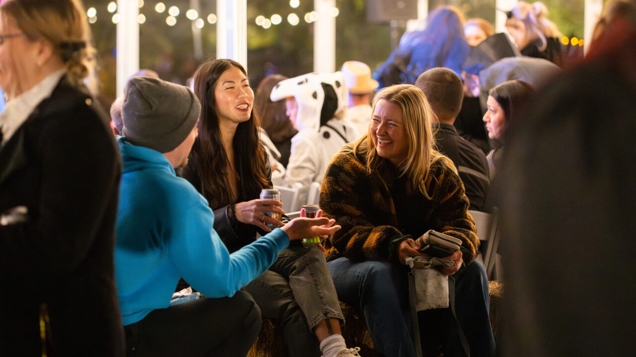 People laughing and chatting at an outdoor evening gathering, surrounded by soft lights