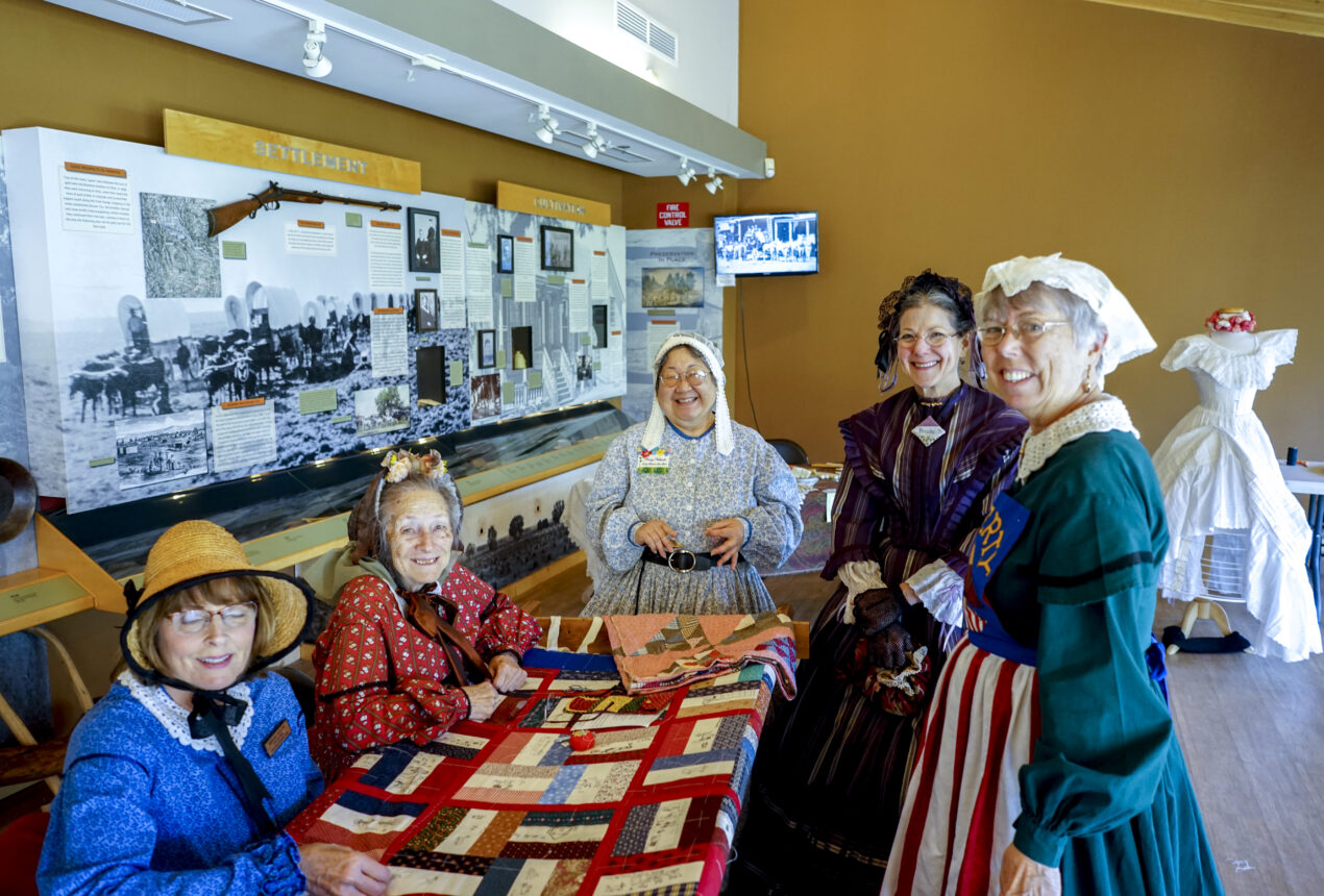 Women in historical clothing smiling around a colorful quilt in a museum exhibit on settlement