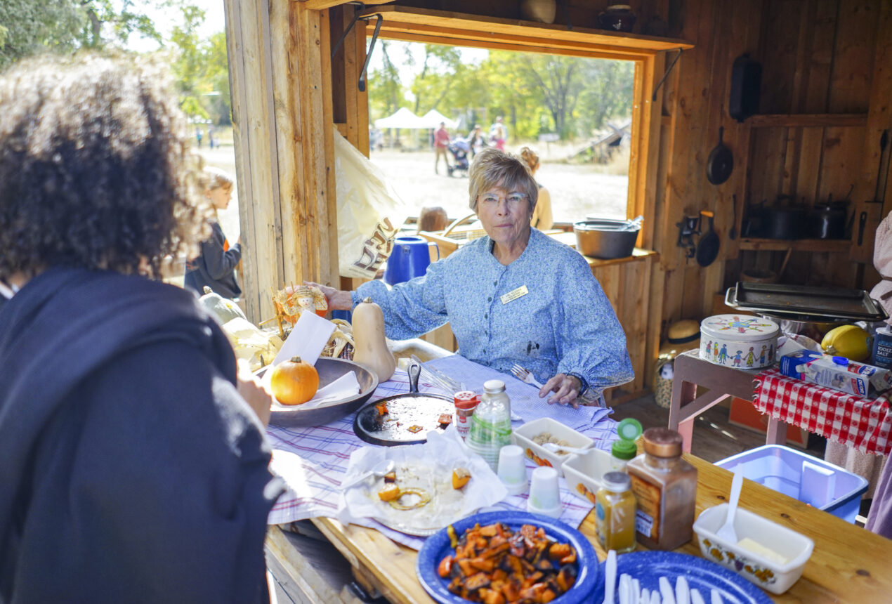 Older woman in a blue dress serving food at a rustic outdoor food stand