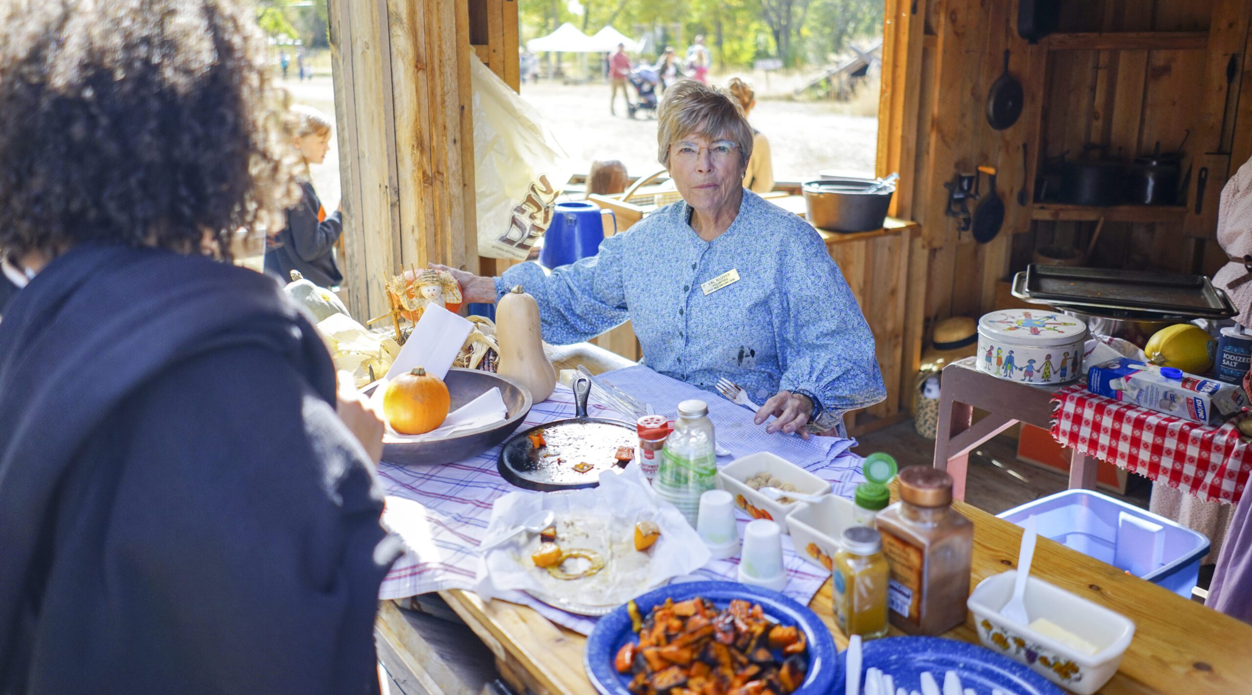 Person in period clothing at rustic outdoor food station with pumpkins and cookware