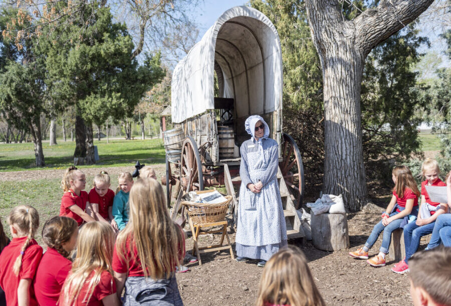 Person in prairie dress stands by a covered wagon, speaking to children sitting in a circle outdoors
