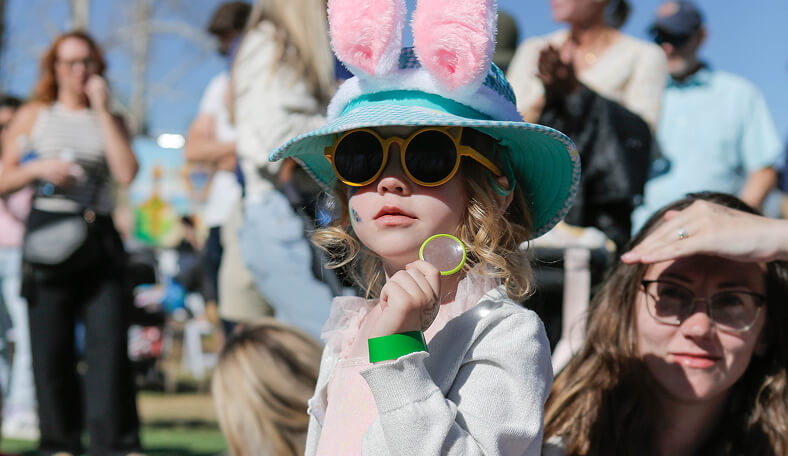 Child in sunglasses and bunny ears at outdoor event, holding a small object, with crowd in background