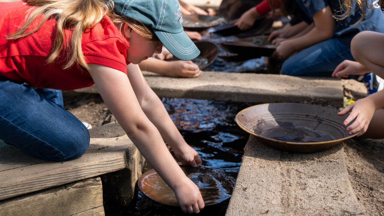 Child wearing a cap pans for gold in a creek with others nearby