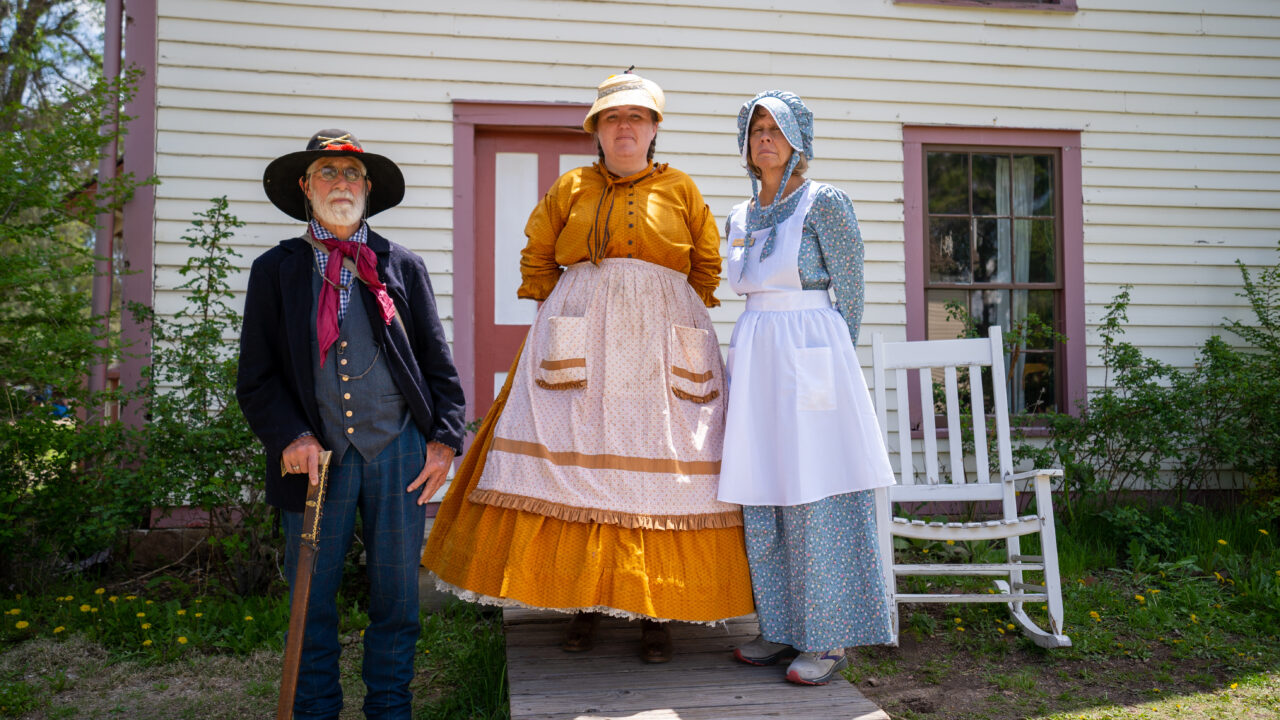 Three people in period clothing stand by a wooden house and rocking chair